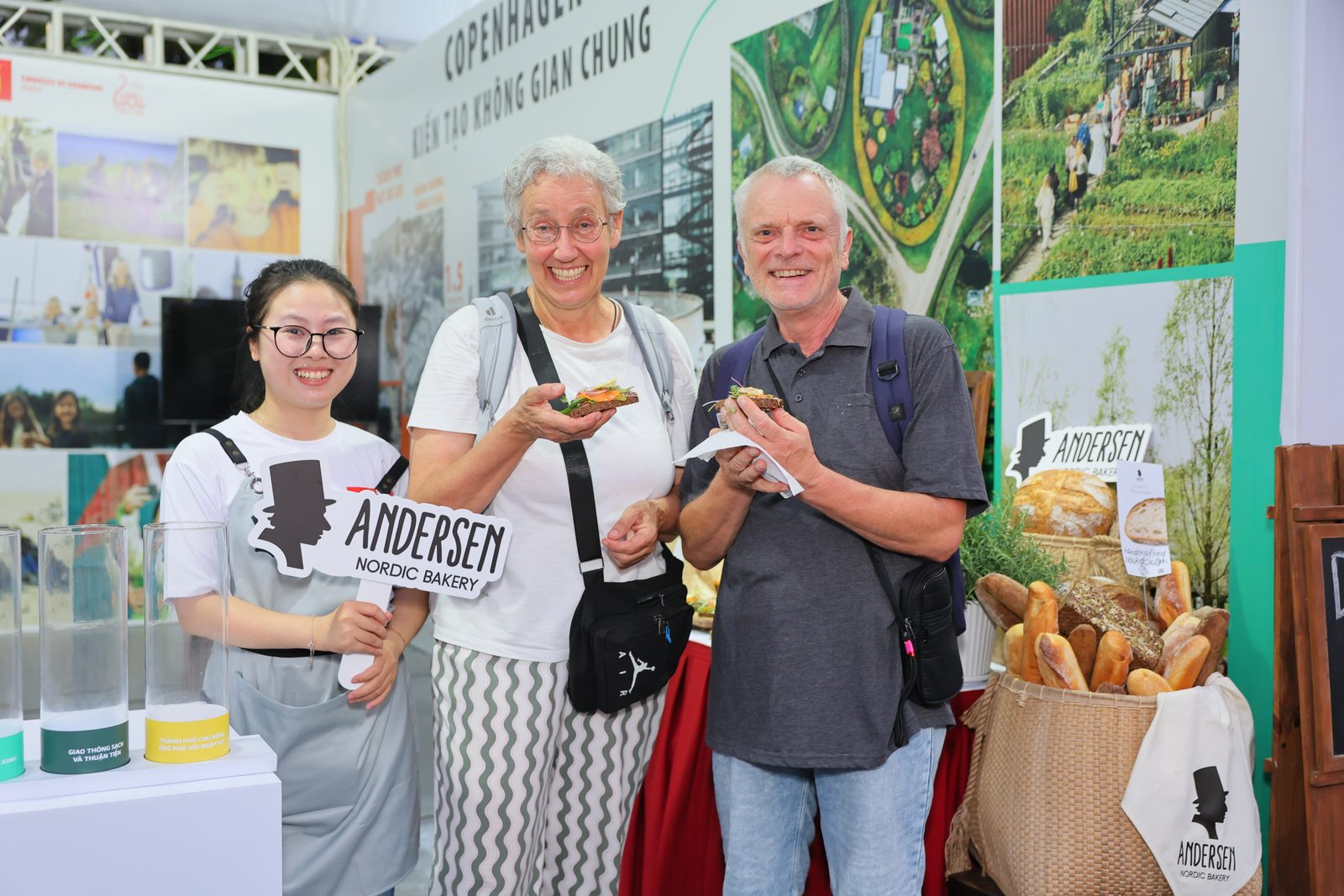 Two visitors hold open sandwiches next to an Andersen team member and a basket of bread